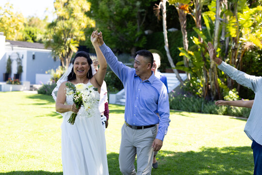 Celebrating senior wedding, diverse couple holding hands and smiling with guests in garden