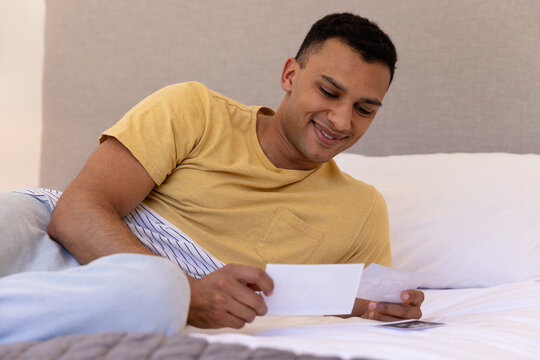 Smiling man relaxing on bed, looking at photographs, feeling nostalgic, at home
