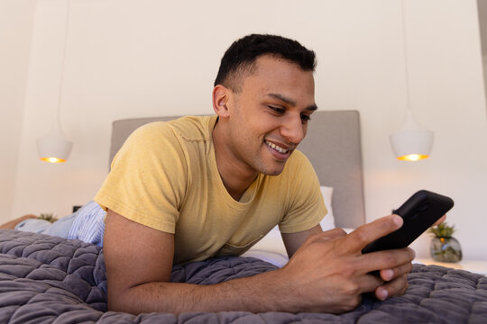 Smiling man lying on bed using smartphone at home, enjoying leisure time