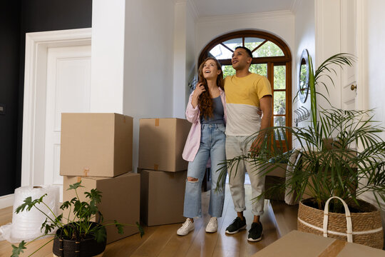 Young diverse couple standing in new home surrounded by moving boxes, looking excited