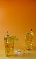 Two glasses of a ice tea with lime and lemon slices in them. The drink is served in a glass with mint leaves on the top and white and red straws. Three ice cubes on the side. Warm orange background.