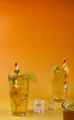 Two glasses of a ice tea with lime and lemon slices in them. The drink is served in a glass with mint leaves on the top and white and red straws. Three ice cubes on the side. Glass of ice tea in the b