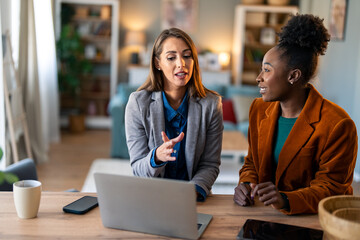 Two Diverse Business Women Having Productive Discussion At Home Office Desk With Laptop And Coffee