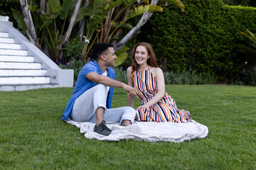 Young diverse couple sitting on blanket in garden, enjoying conversation and laughter