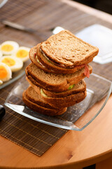 A photo of stacked breakfast sandwiches made with cucumber, cheese, and salami on whole wheat bread, with defocused boiled eggs in the background