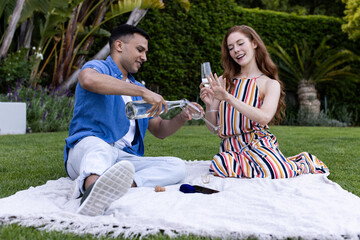 Young diverse couple enjoying picnic on grass, pouring sparkling water into glasses, in garden