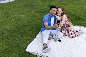 Showing engagement ring, young diverse couple on picnic blanket, looking at smartphone, in garden