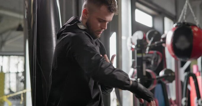 A young man in a dark tracksuit wraps bandages around his hands before a boxing match or other fighting competition or training