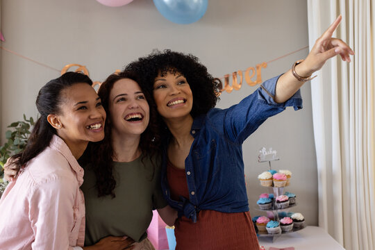 Diverse female friends at baby shower, smiling and pointing with cupcakes in background, at home