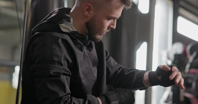 A young man in a dark tracksuit wraps bandages around his hands before a boxing match or other fighting competition or training