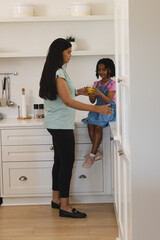 Mother giving juice to daughter sitting on kitchen counter, both smiling warmly