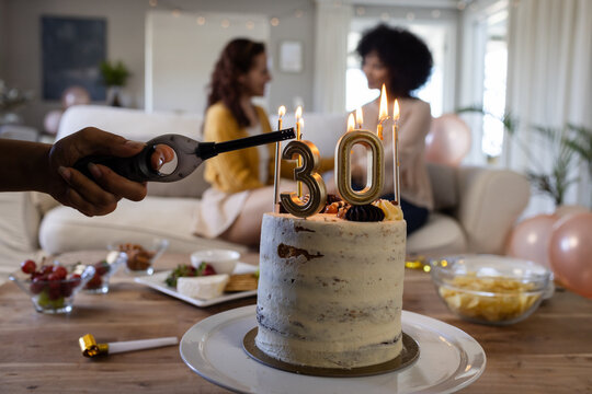 Lighting candles on birthday cake while diverse female friends chat on couch at home
