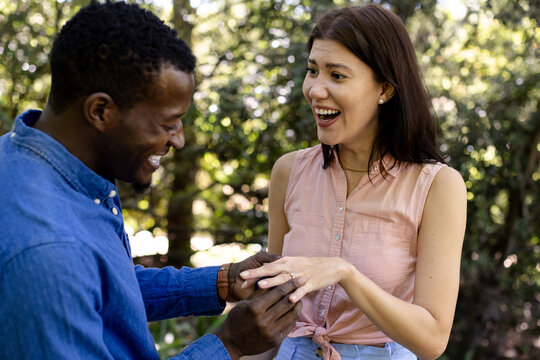 Man proposing to surprised woman outdoors, placing ring on her finger, smiling