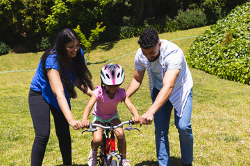 Parents teaching daughter to ride bicycle in sunny park, smiling and supportive