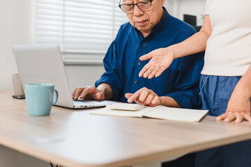 Obraz premium Elderly man reviews pension savings on laptop while woman gestures supportively beside him.