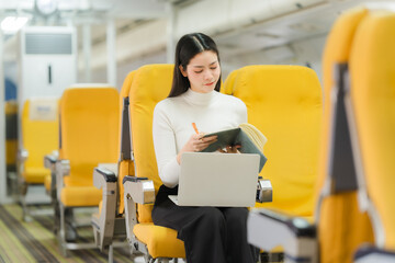 Focused Traveler in Train: A focused Asian woman works on a laptop while taking notes on a notebook...