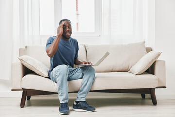 An emotional young African American man in casual clothing sits on a sofa, frustrated while using a laptop in a bright, minimalistic room