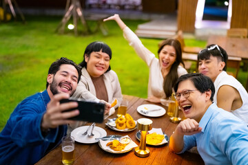 Group of Happy Asian millennial people friends enjoy and fun celebration reunion meeting using mobile phone taking picture together at dinner party in the garden at restaurant on holiday vacation.