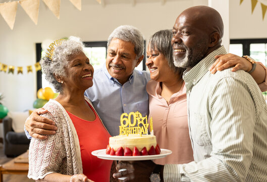 Celebrating 60th birthday, diverse senior friends hugging and smiling with cake at home - Powered by Adobe