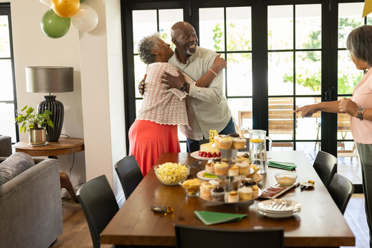 Senior diverse senior couple hugging joyfully at home during party celebration with friends - Powered by Adobe