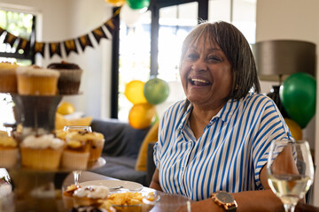 Senior woman laughing at party table with cupcakes and decorations, at home