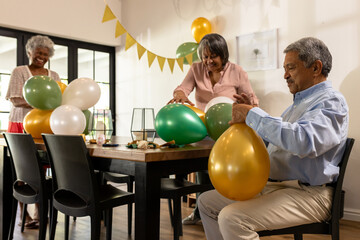 Diverse senior friends preparing for celebration, inflating colorful balloons around table, at home