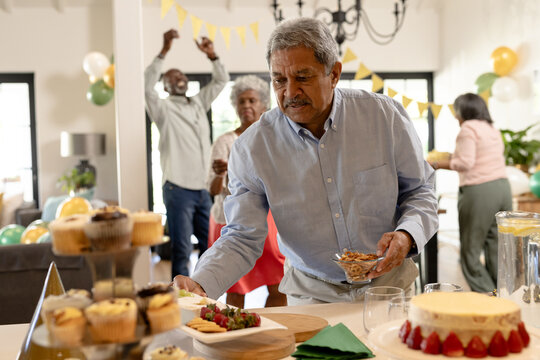 Celebrating with diverse senior friends, man savoring dessert table while guests cheer, at home