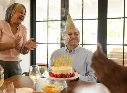 Celebrating 60th birthday, senior man with cake and diverse senior friends applauding, at home