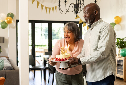 Smiling diverse senior couple holding birthday cake with candles in festive living room, at home