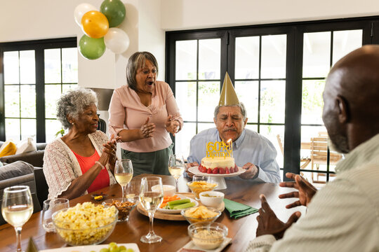 Diverse senior friends celebrating birthday at home, senior man blowing out candles on cake