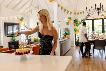 Blonde woman celebrating 21st birthday, lighting candles on cake in decorated home