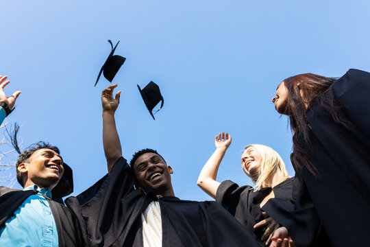 Diverse graduates celebrating by throwing caps in air, smiling under clear blue sky, copy space