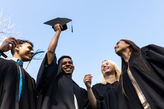 Diverse graduates celebrating in garden, smiling and holding caps under clear blue sky