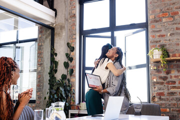 Diverse female colleagues celebrating success with congratulatory hug and award in office