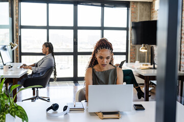 Focused diverse female professionals working on laptops in modern office, collaborating on projects