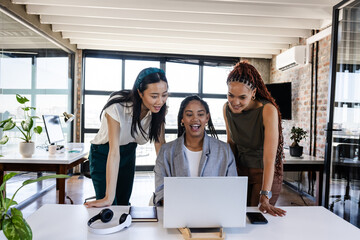 Diverse businesswomen celebrating success while looking at laptop in modern office