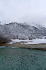 Kranjska Gora's winter beauty unfolds with a clear river reflecting the snowy landscape, frosted trees, and mist-shrouded Julian Alps, capturing a serene Slovenian scene.