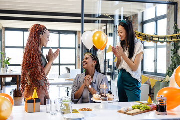 Female diverse colleagues celebrating birthday in office with cake and balloons, smiling happily