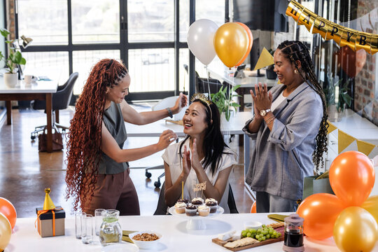 Female diverse colleagues celebrating birthday in office with cake and balloons, smiling joyfully