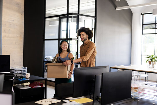 Carrying box of office supplies, diverse colleagues smiling walking through modern workspace