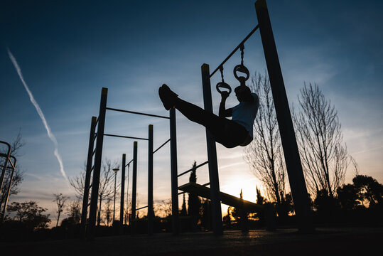 young athletic man exercising fitness calisthenics workout and making ring leg raises for abdomen abs muscles at street gym during sunset, body improvement and strength training