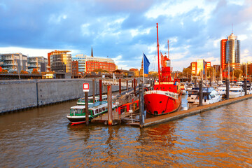 Obraz premium Hamburg waterfront scene with red ship anchored beside other vessels, with modern architecture and a dramatic sky forming perfect backdrop. Harbor scene in Hamburg Germany with variety of boats docked