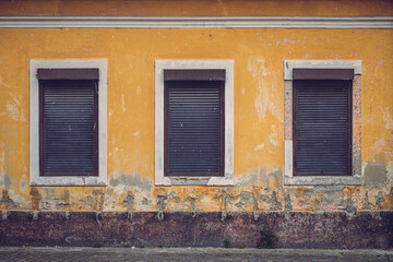 A weathered yellow wall with three closed shutters creates a striking geometric composition. The peeling paint and textured decay evoke a sense of time, history, and forgotten stories