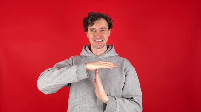 young adult curly haired brunette man holds his hands in front of shows sign timeout gesture. smiling, wearing grey hoodie. isolated on red studio background. Concept - pause, break, rest, vacation