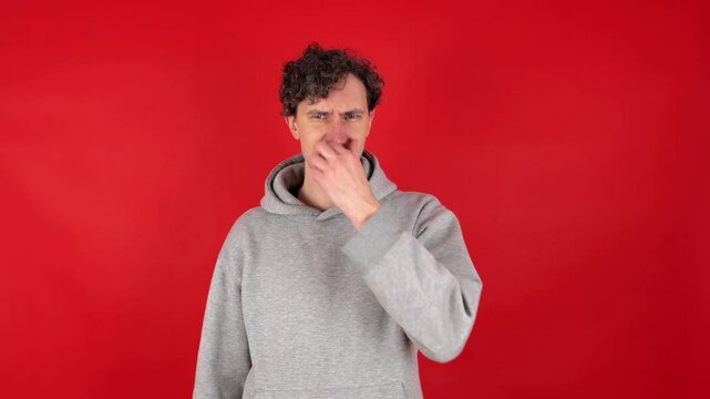 Portrait displeased unhappy young adult curly man squeezing nose fingers, waves hand in front reacting gross, smelly odor. Isolated on red background studio shot. Capturing strong reaction to smell.
