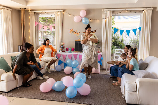 Diverse female friends celebrating baby shower with balloons and gifts in cozy living room, at home