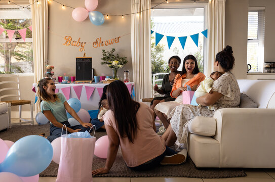 Diverse female friends enjoying baby shower with balloons and gifts in living room, at home - Powered by Adobe