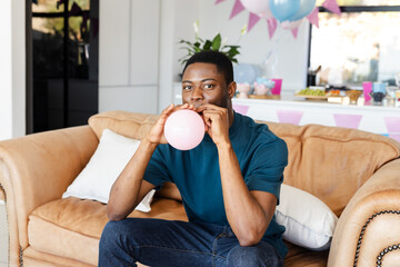 Baby shower, African American man blowing up balloon on couch at home party, looking cheerful