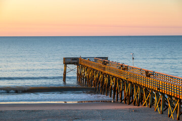 Wooden pier at seashore, showing beach and horizon over water, photographed at golden hour of morning.