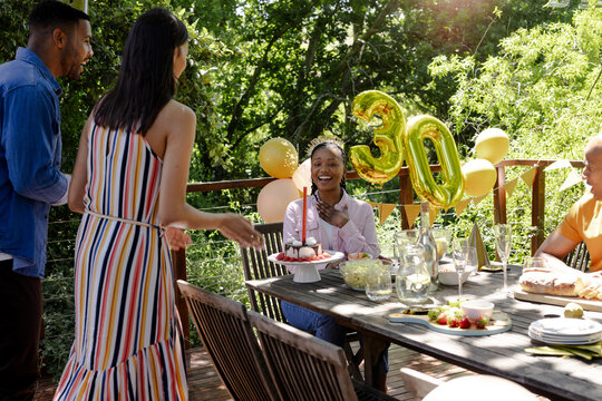Celebrating birthday outdoors in garden, African American woman smiling with diverse friends and cak